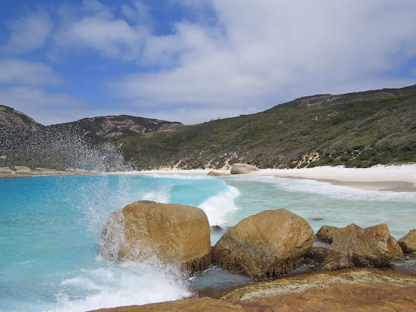 Clear blue water crashing into a granite rock with a beautiful white sandy beach behind.
